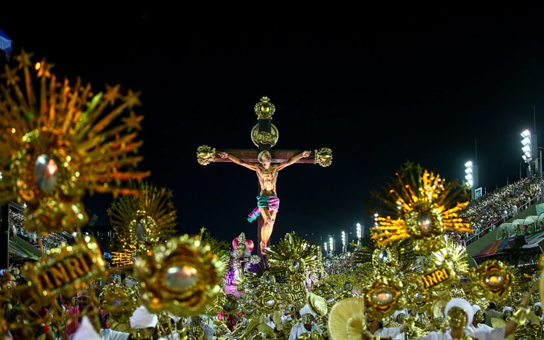 Multidão em desfile de Carnaval com símbolo religioso em alegoria, gerando debate.