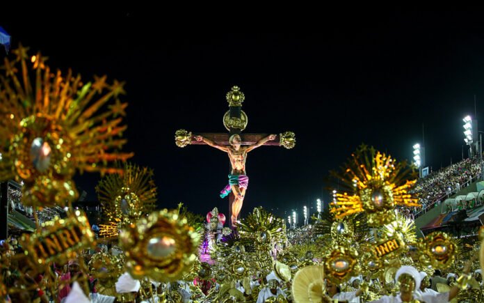Multidão em desfile de Carnaval com símbolo religioso em alegoria, gerando debate.