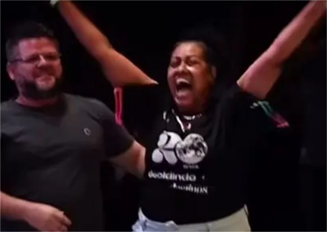 Mulher chorando de emoção e gratidão com os braços erguidos em um palco de evento religioso após um testemunho de cura.