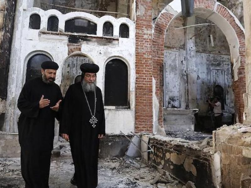 Rua de vilarejo egípcio com tensão após ataque de multidão, mostrando sinais de vandalismo e observadores apreensivos.