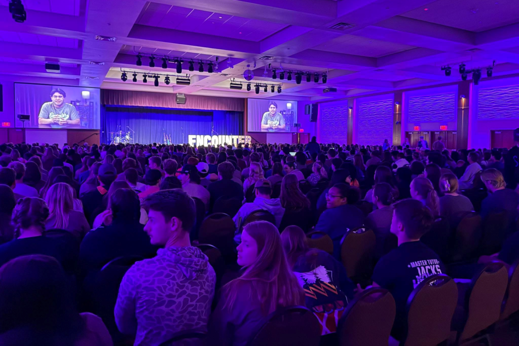 Jovens estudantes em um evento religioso lotado em uma universidade americana, com expressões de fervor e atenção.