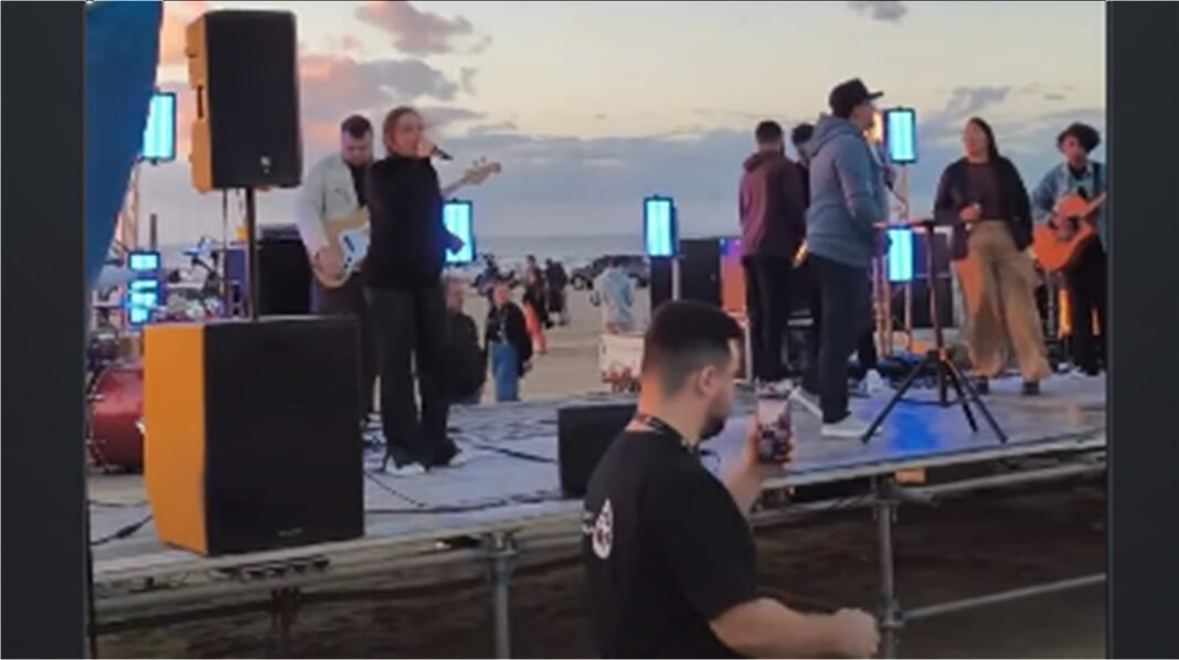 Multidão reunida em praia para culto ao nascer do sol com cânticos e pregação.