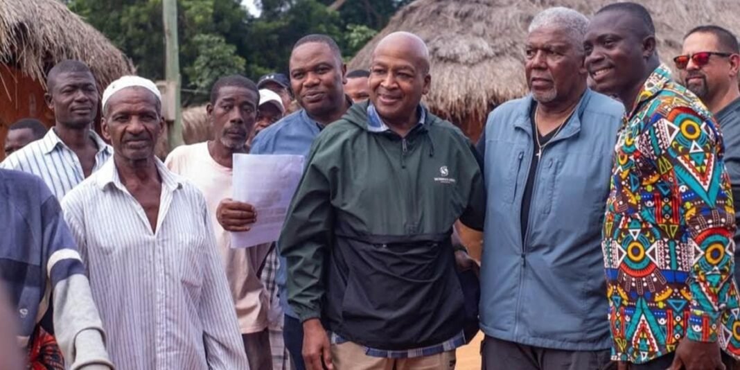 Pastor Godwin K. Ahlijah sorrindo ao lado de um poço de água em Gana, com crianças e moradores ao fundo.