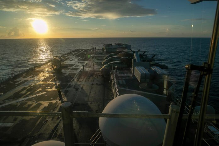 Navio de guerra USS Tripoli navega em águas do Golfo Pérsico com a Ilha Kharg ao fundo.