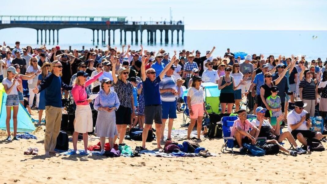 Pessoas reunidas em uma praia para batismos em massa, com cerimônias ocorrendo na água e espectadores na areia.