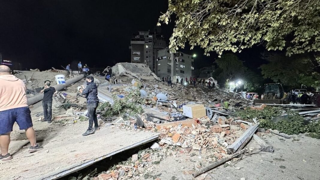 Rua em Caracas coberta de escombros após terremoto, com pessoas em choque observando prédios destruídos.