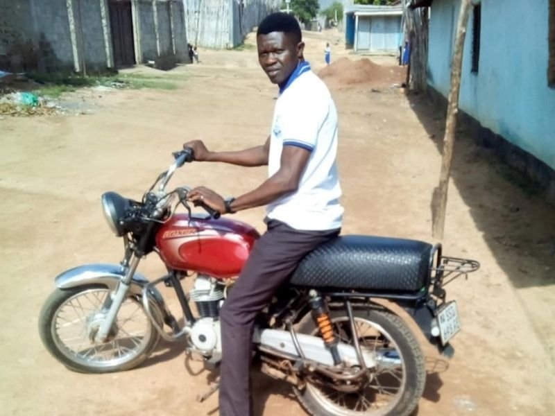 Jovem refugiado sudanês sorri ao lado de uma motocicleta que o ajuda a recomeçar a vida.