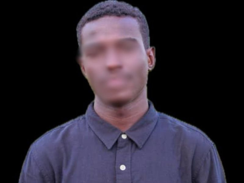 Jovem sudanês atrás das grades de uma prisão, olhando para fora com expressão aflita.